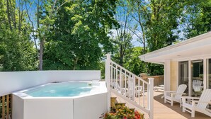 Un jacuzzi en una terraza de madera con barandas y sillas blancas, rodeado de exuberante follaje.