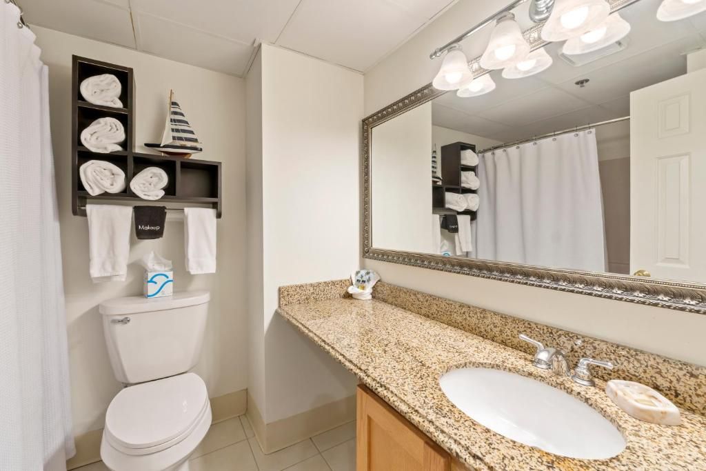 Une salle de bain avec des toilettes blanches, un lavabo sur un plan de travail en granit, un grand miroir et une étagère avec des serviettes pliées.