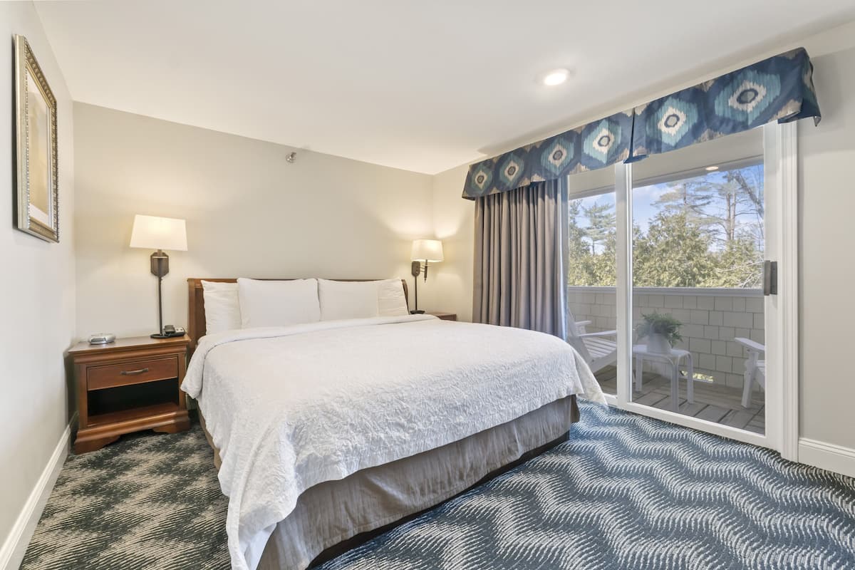 A bedroom with a large bed, two bedside tables with lamps, a patterned rug, and a view through a sliding glass door to an outdoor patio.