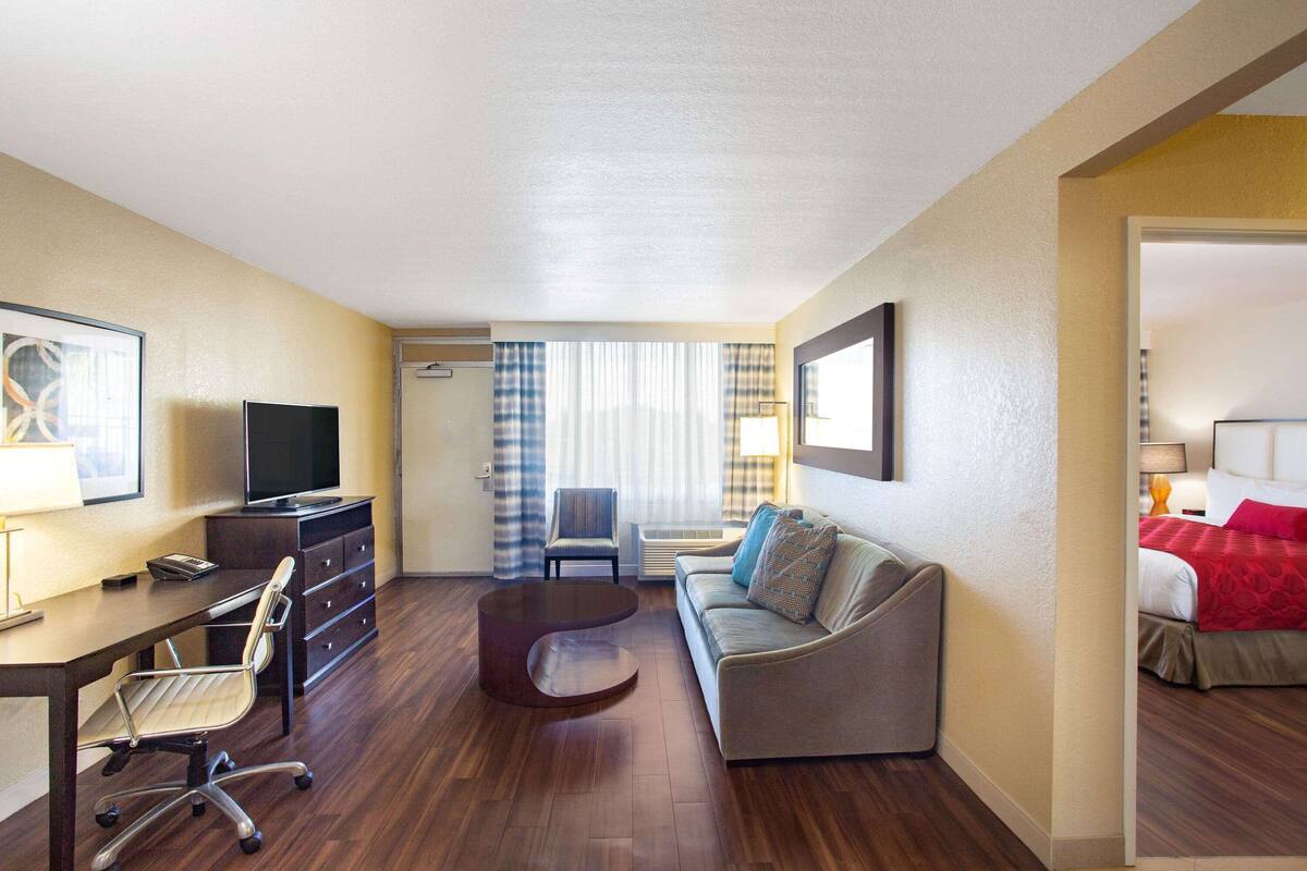 Habitación de hotel con piso de madera, un escritorio con silla, un sofá, un televisor y una cama con ropa de cama roja.