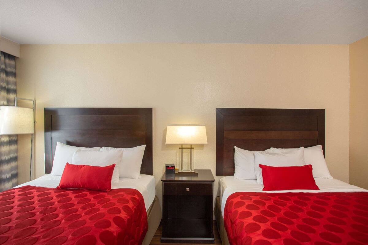 Habitación de hotel con dos camas, cada una con una colcha roja de lunares, almohadas blancas y cabeceros de madera oscura. Entre las camas hay una mesita de noche con una lámpara y un pequeño reloj.