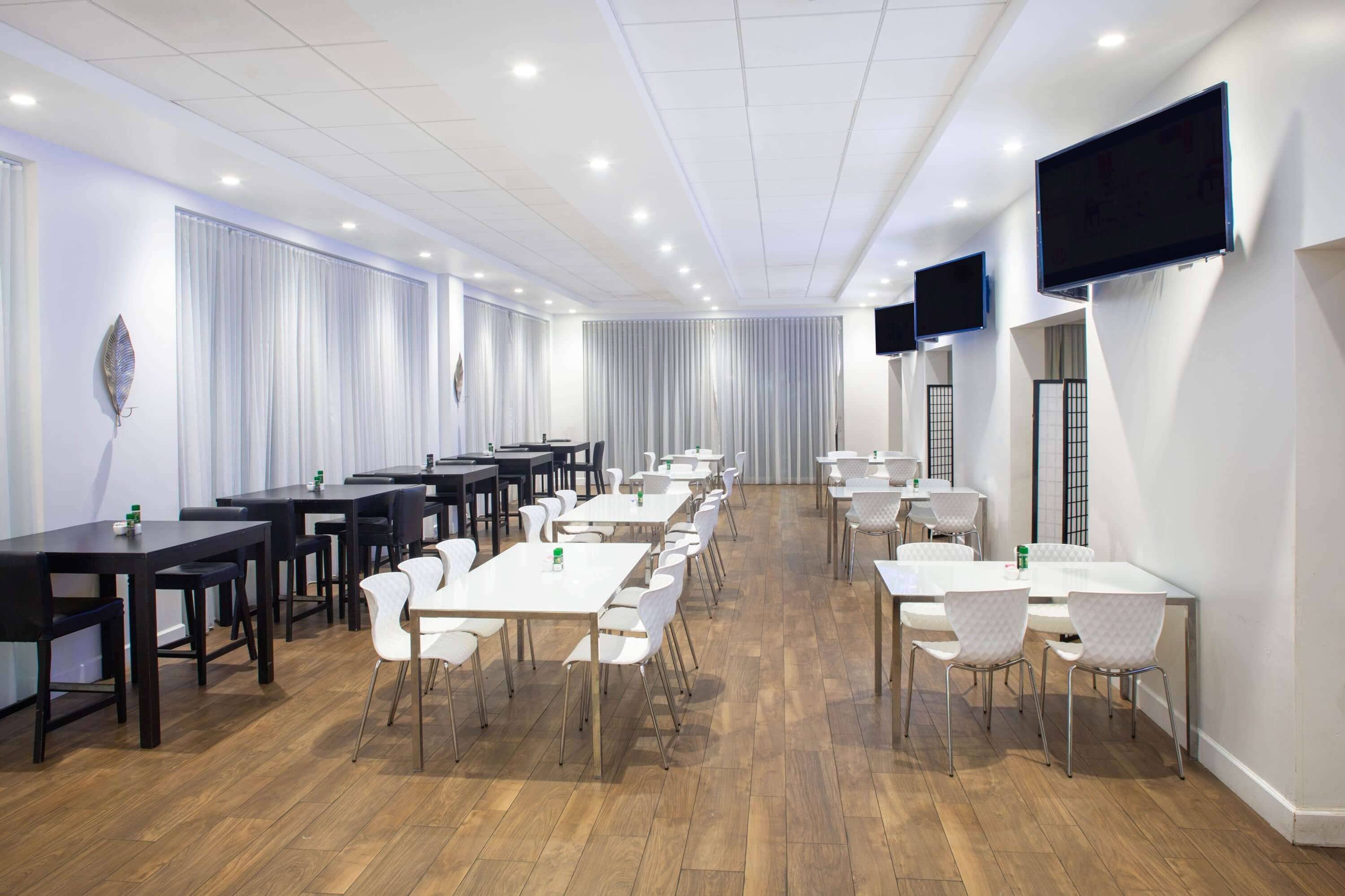 A conference room with multiple tables and chairs, white curtains, and mounted televisions on the walls.