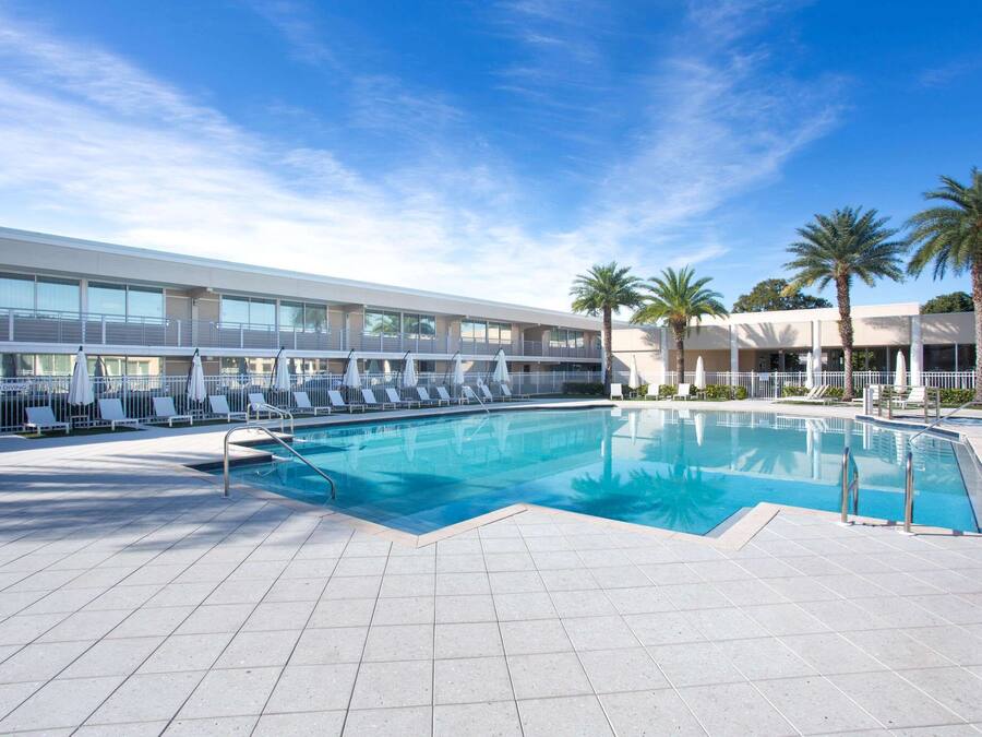 A swimming pool with lounge chairs and umbrellas, surrounded by a building with large windows and palm trees.