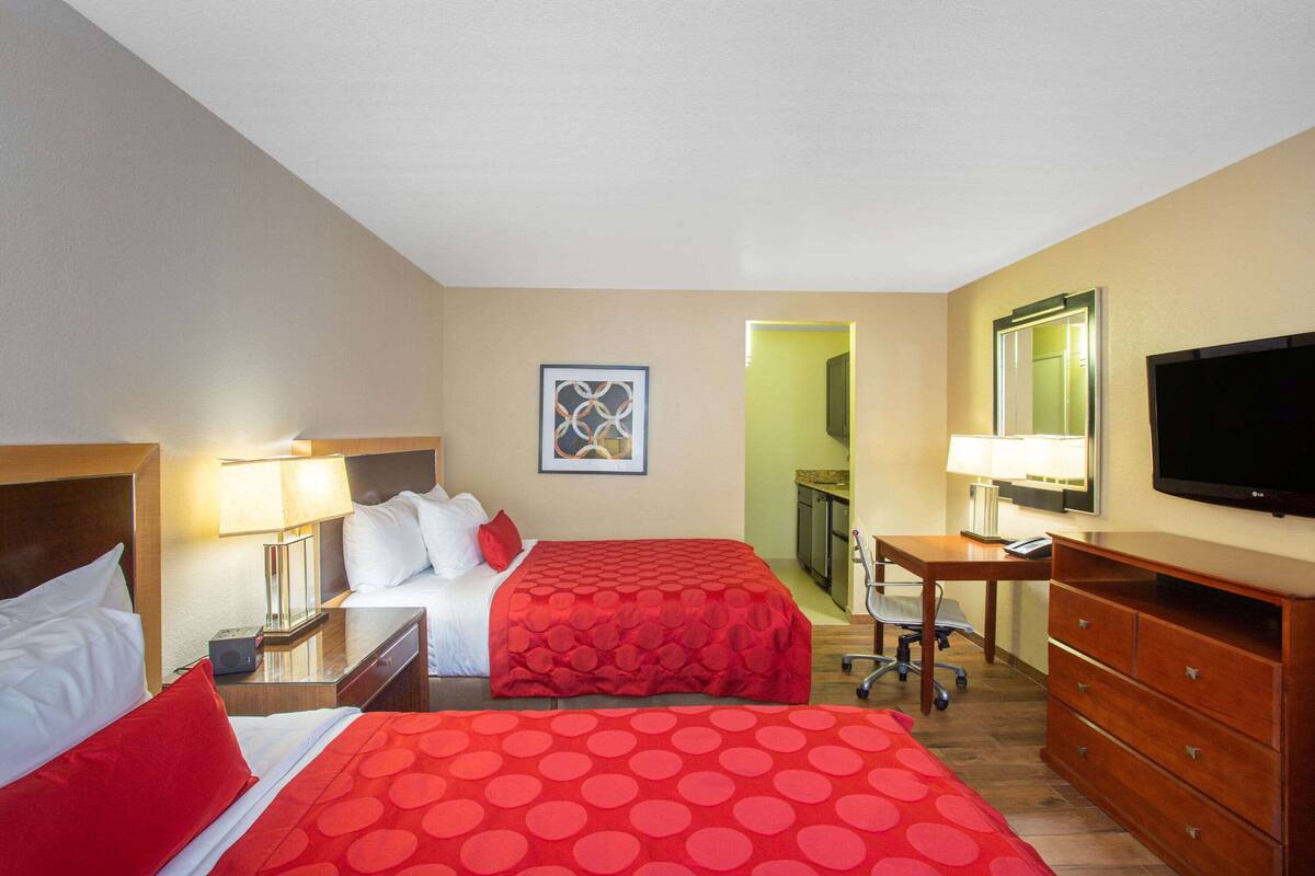 Habitación de hotel con dos camas, un escritorio, una silla, un televisor y un baño visible a través de una puerta abierta.