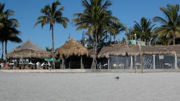 Zona de playa con construcciones de techo de paja, palmeras y un primer plano arenoso.