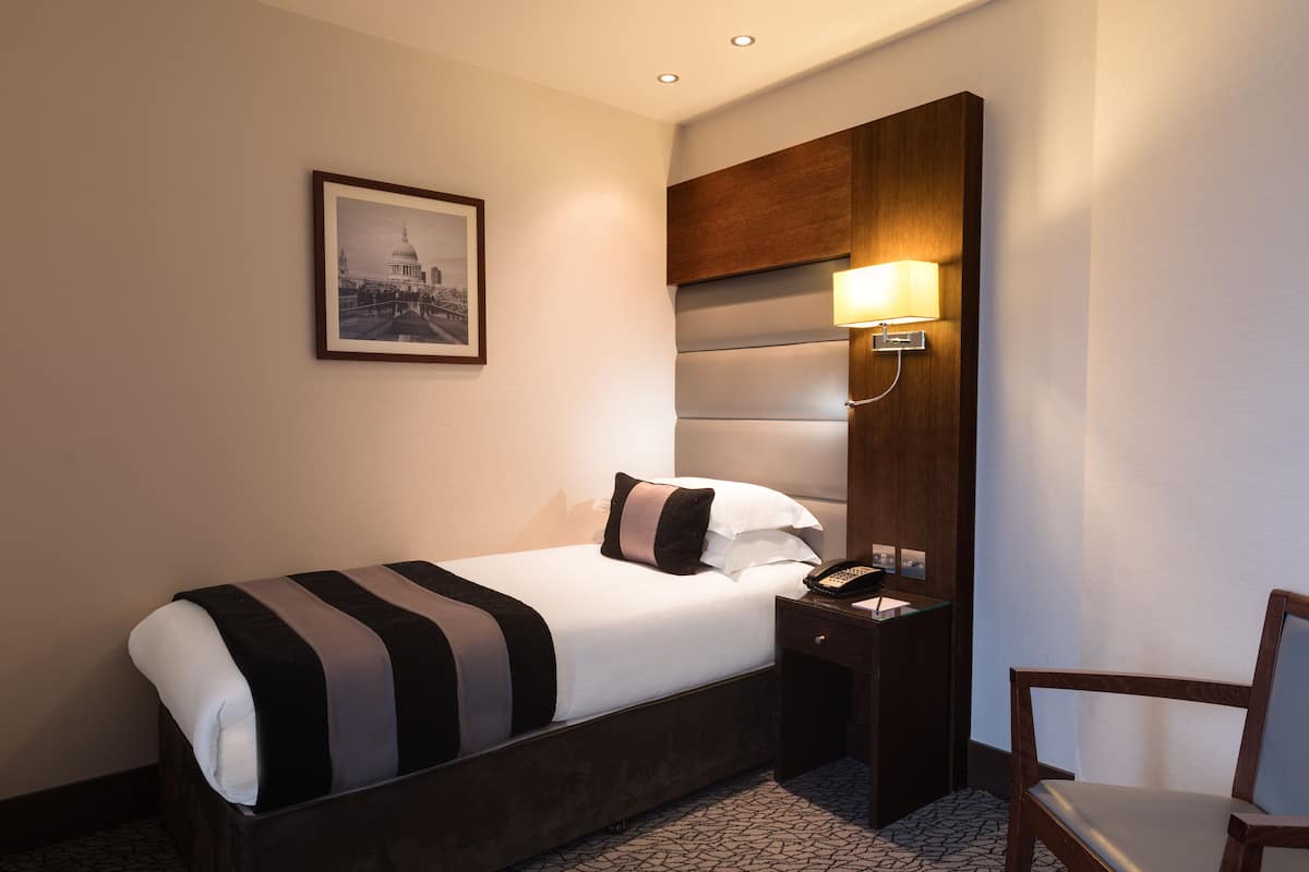 Een hotelkamer met een bed, een nachtkastje met een lamp, een stoel en een ingelijst schilderij aan de muur.