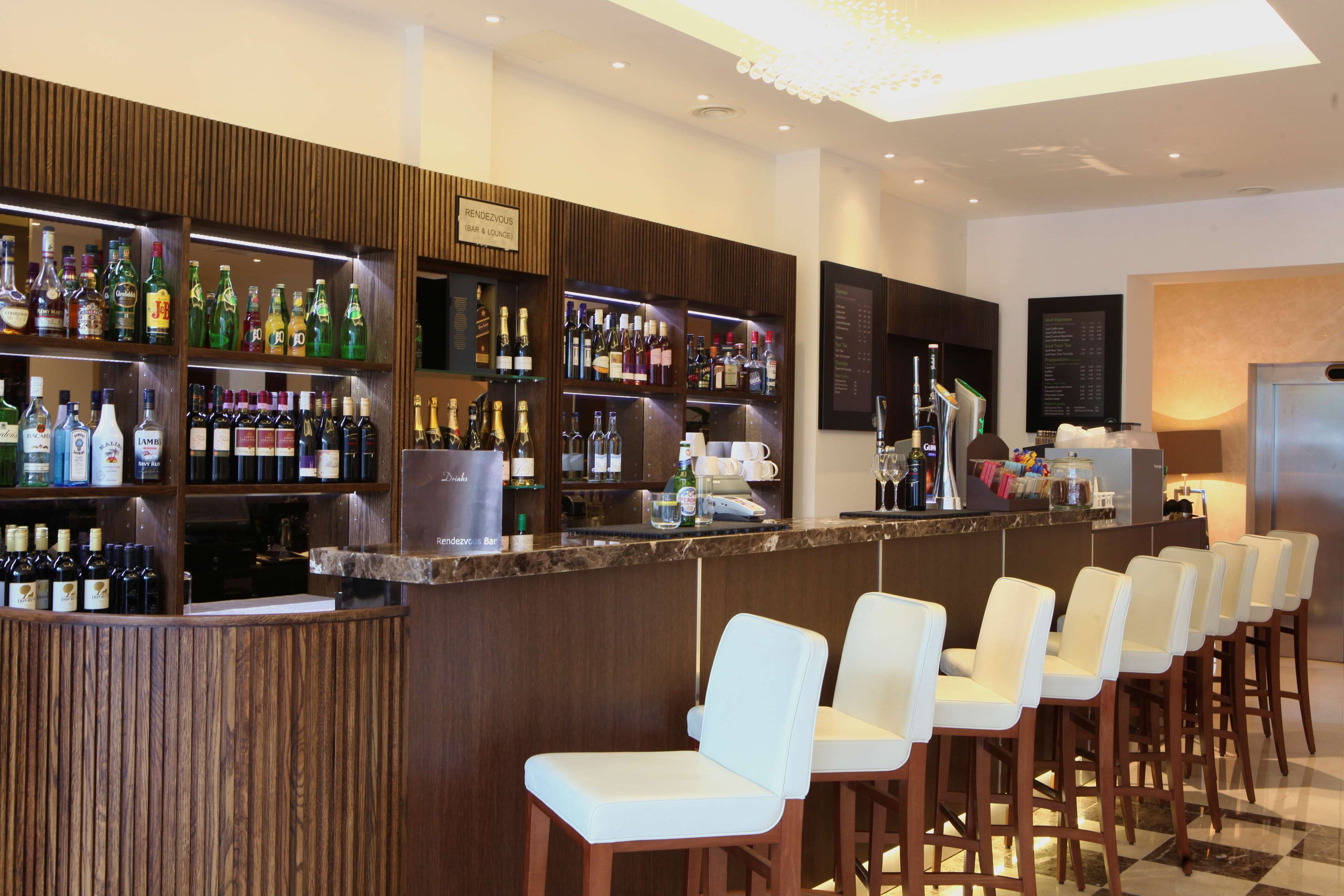 A bar area with multiple shelves of liquor bottles, a bar counter, and several white bar stools.