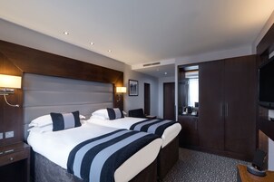 Habitación de hotel con dos camas, un escritorio, un televisor y un baño visible a través de una puerta abierta.