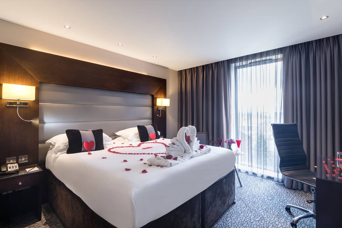 Een hotelkamer met een groot bed, hoofdeinde, nachtlampjes, een bureau met stoel, een raam met gordijnen en een vloerbedekking.