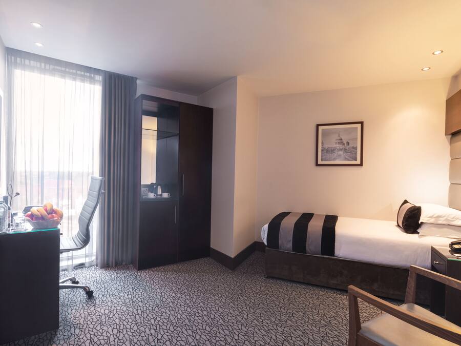 Quarto de hotel com cama, escrivaninha, cadeira, mesa pequena com uma cesta de frutas e um quadro na parede.