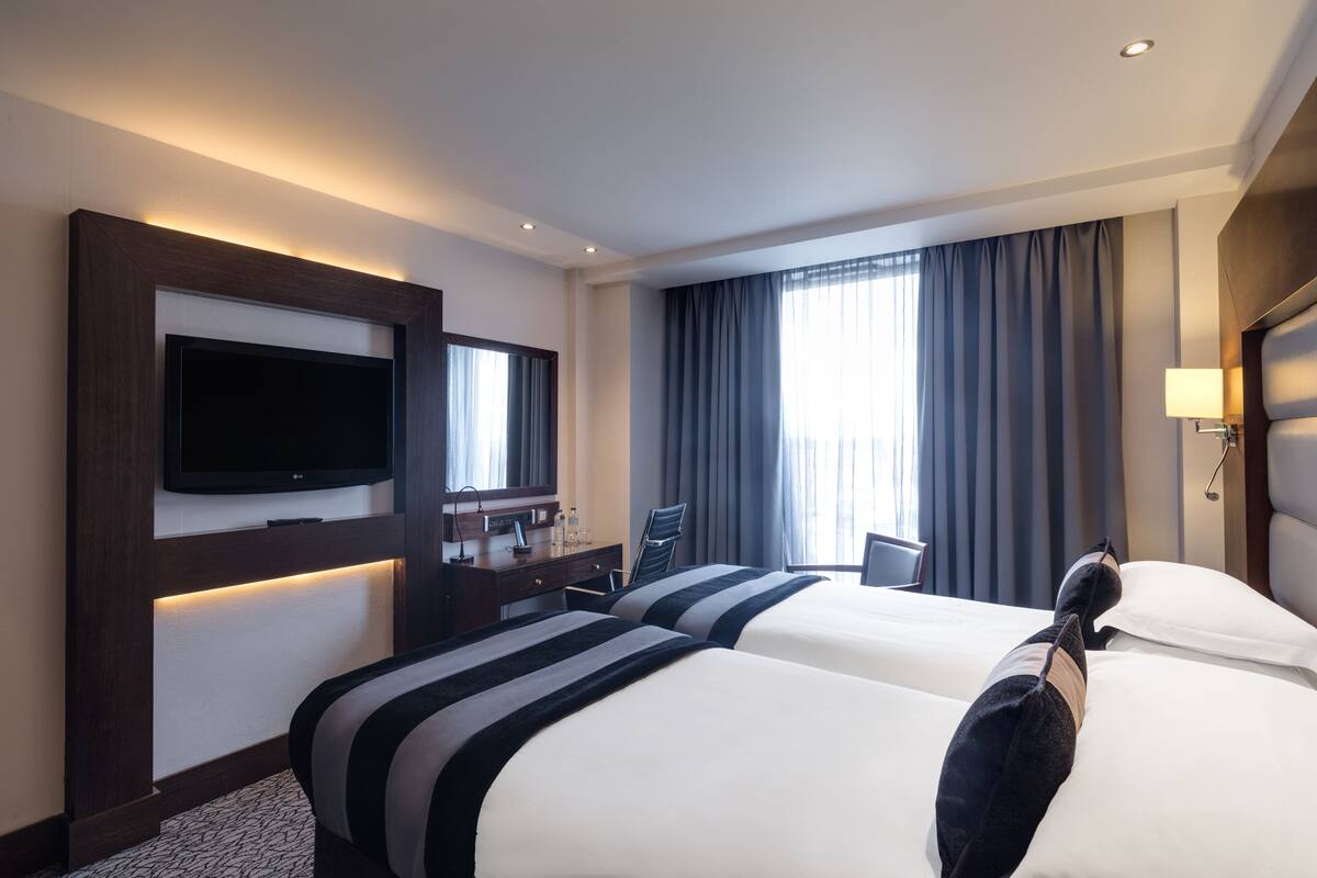Een hotelkamer met twee bedden, een flatscreen-tv, een bureau met lamp en een groot raam met gordijnen.