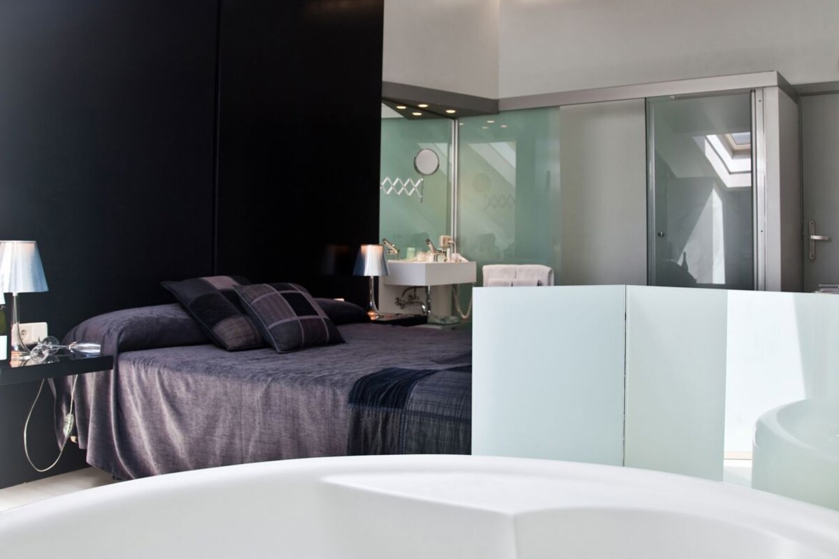 Een moderne slaapkamer met een groot bad, een bed met donkere beddengoed, een nachtkastje met een lamp, en een badkamergedeelte met glazen wanden.