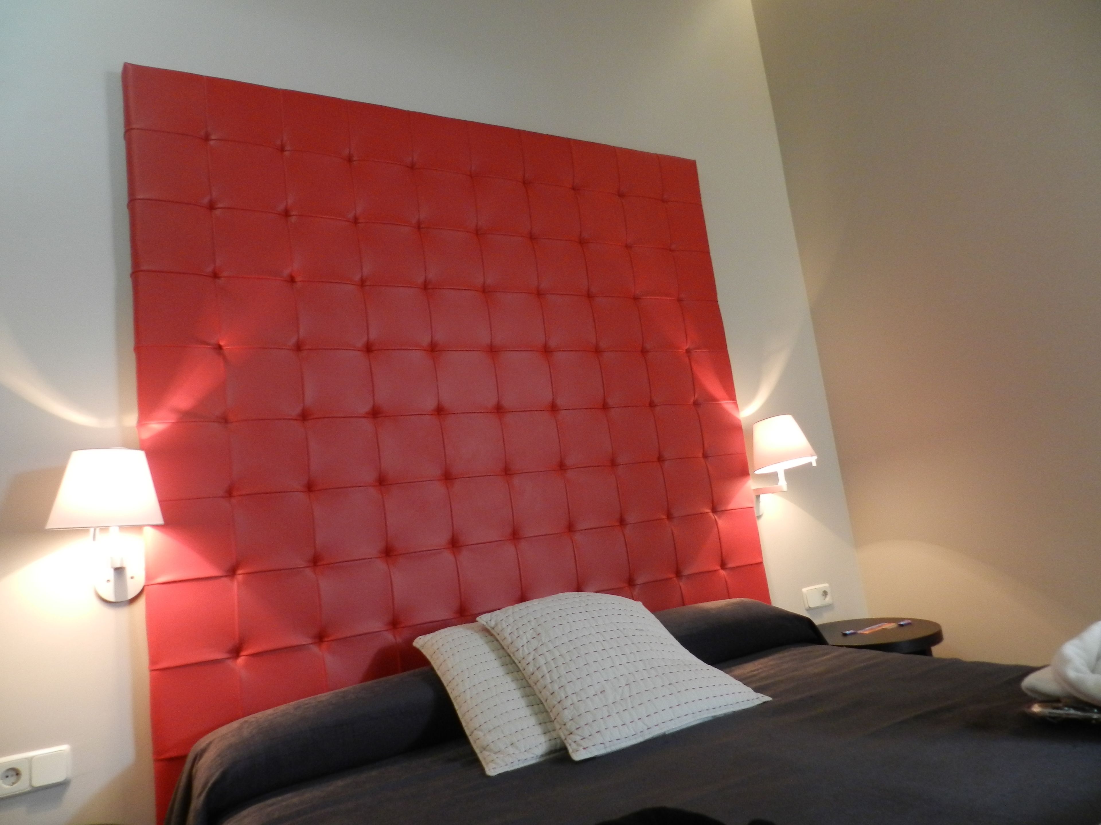 Una habitación de hotel con un cabecero rojo capitonado, dos lámparas de noche, una cama con ropa de cama oscura y una mesita de noche pequeña con un control remoto.