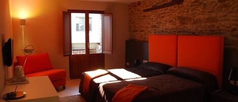 A hotel room with a bed, a desk, a chair, a lamp, and a stone wall.