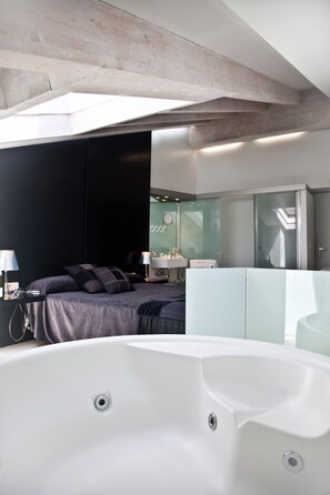 Ein modernes Schlafzimmer mit einer großzügigen Badewanne, einem Bett mit dunkler Bettwäsche, einem Nachttisch mit einer Lampe und einer Duschkabine aus Glas.