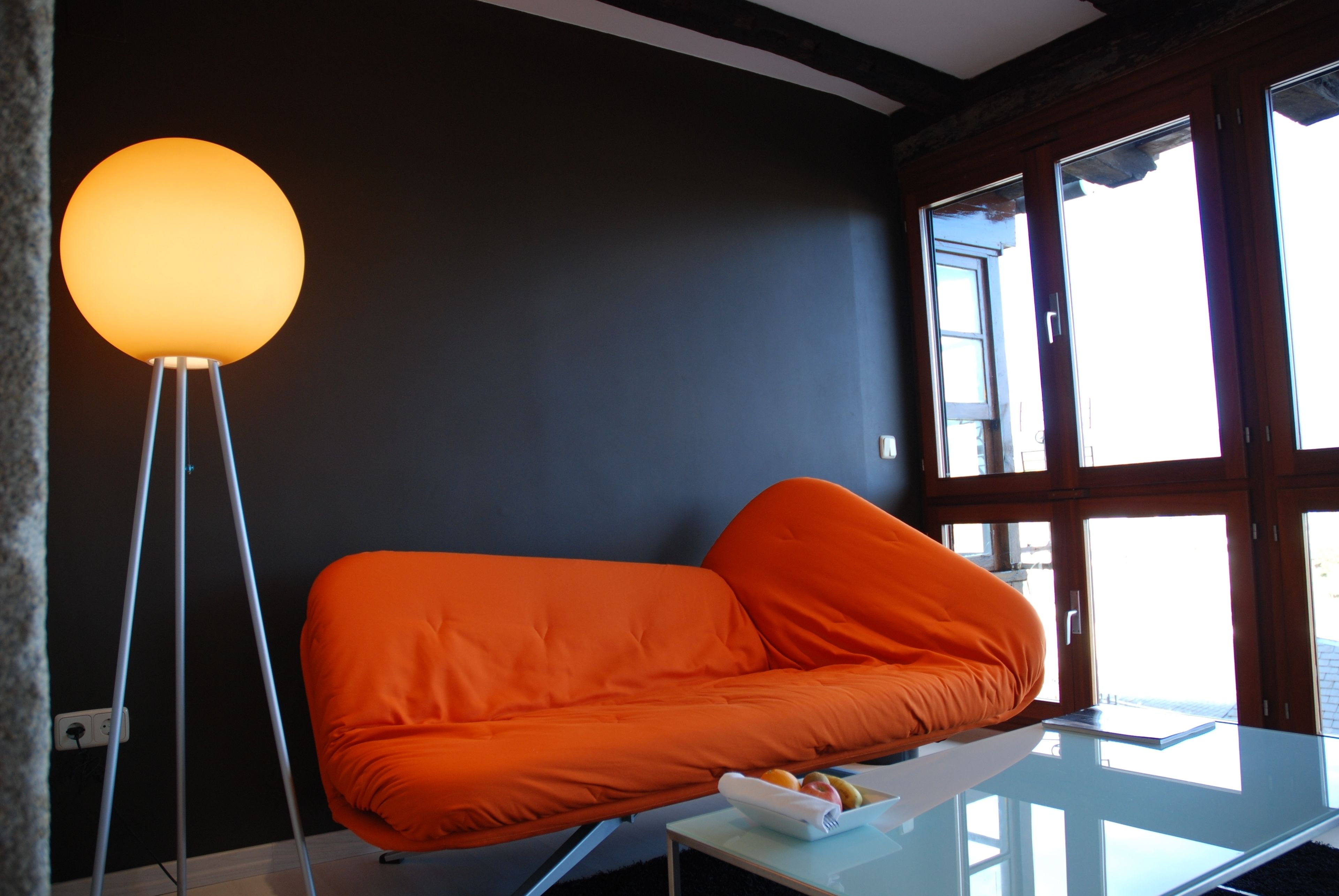 Una habitación moderna con una pared oscura, un sofá naranja brillante, una mesa de centro de cristal y una lámpara de pie.