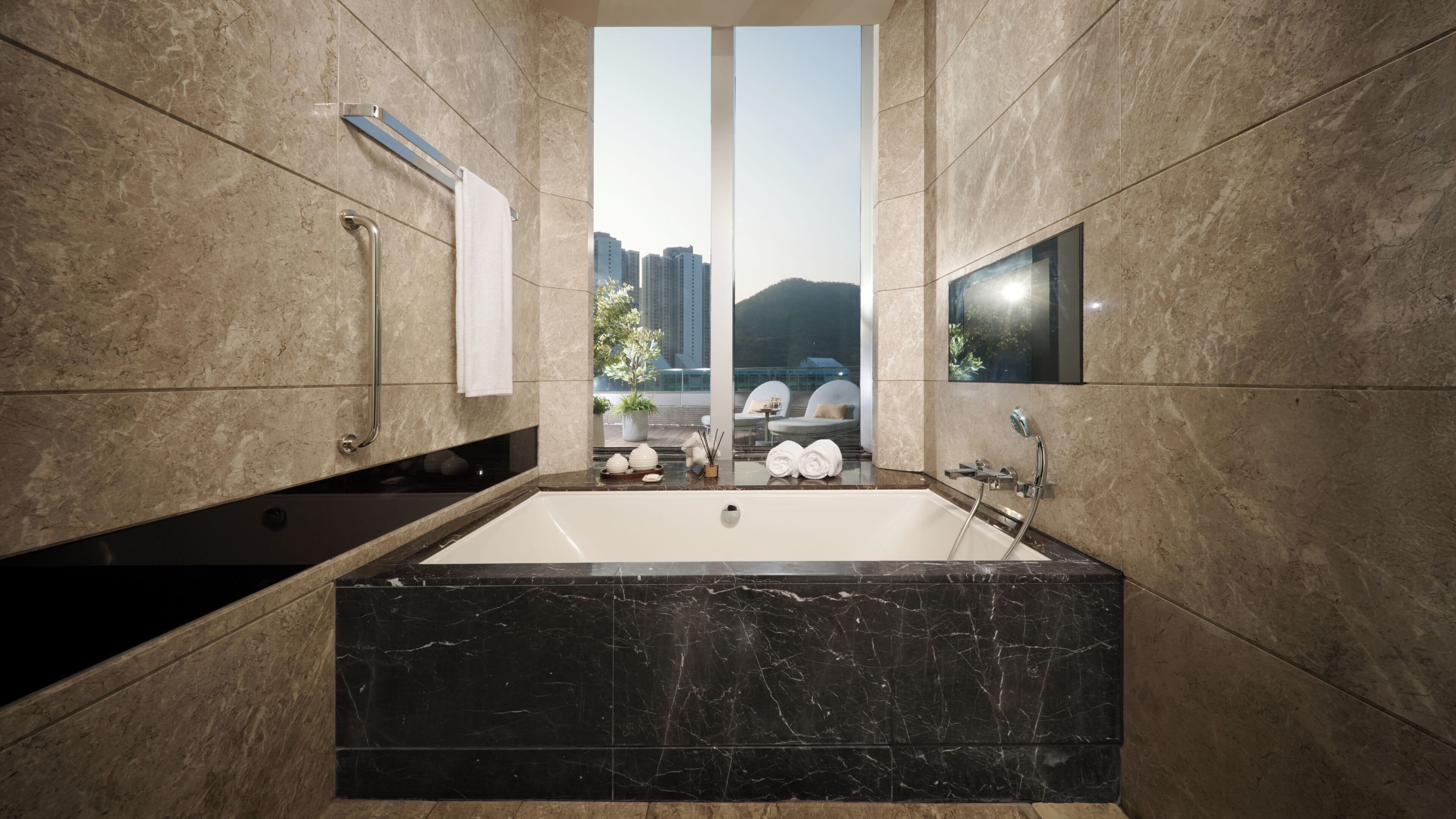 A modern bathroom with a large bathtub, a handrail, a towel rack, and a view of buildings through a large window.