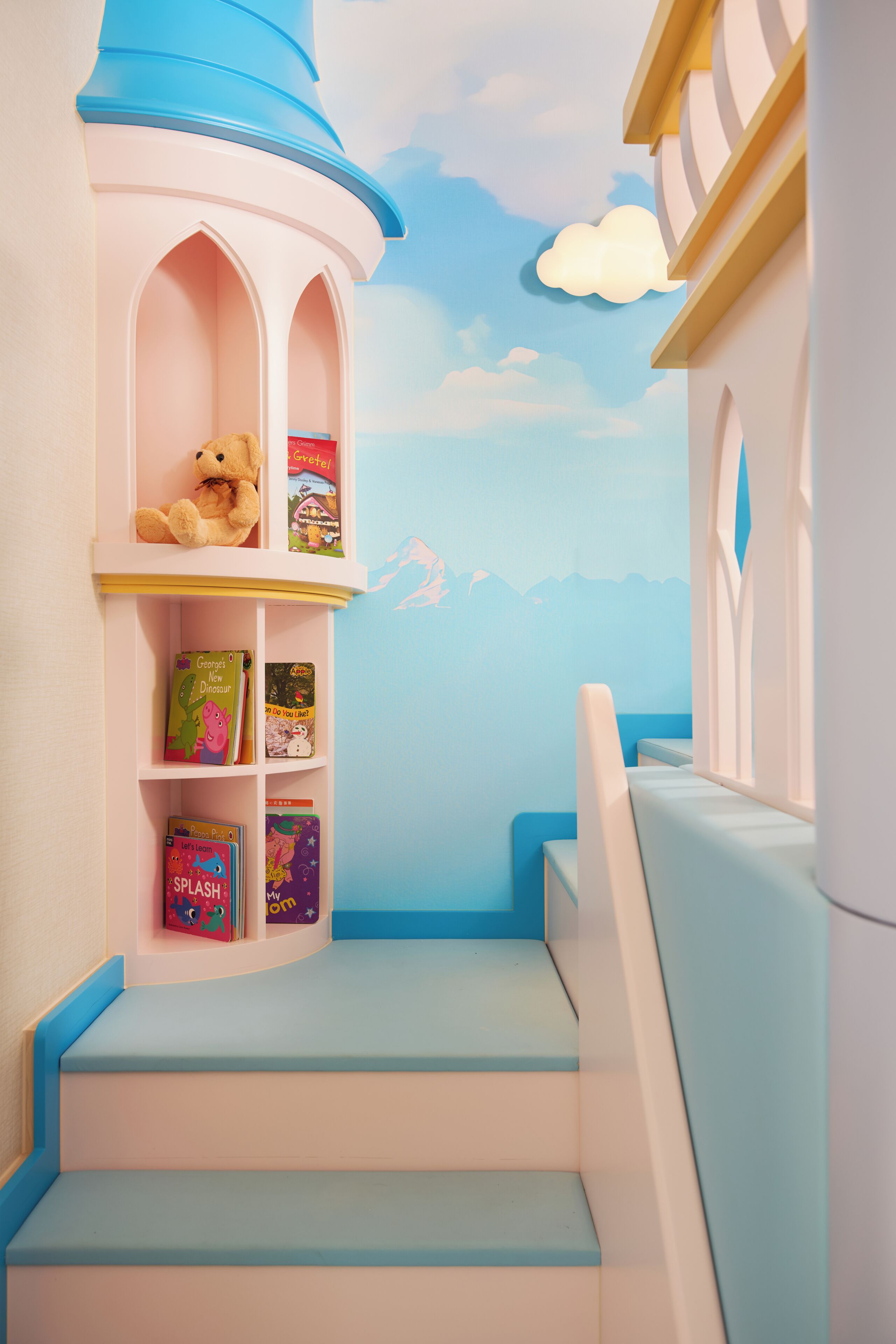 A children's play area with a bookshelf, a teddy bear, and a mural of a sky and clouds.