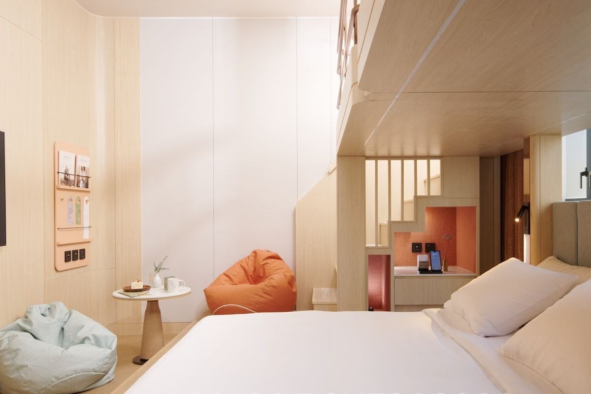 Una habitación de hotel moderna con una cama grande, un televisor, una mesita con una planta y un sillón de peluche.