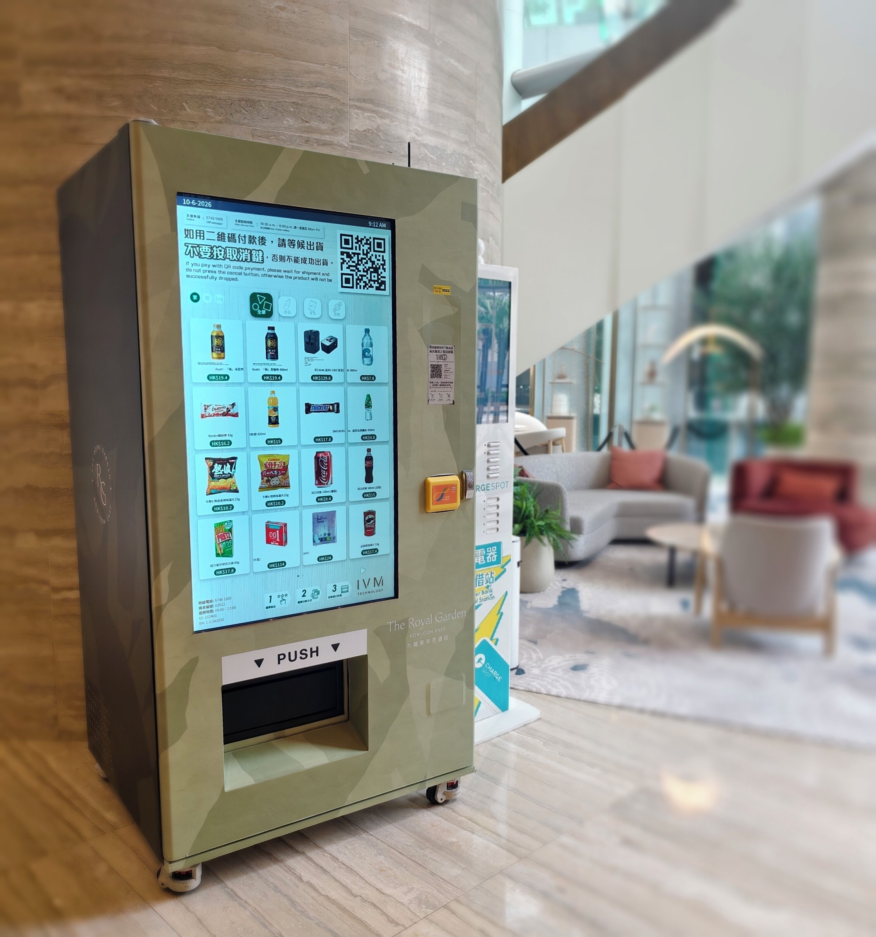 A vending machine with a digital display, located in a modern lobby with a seating area and a plant.