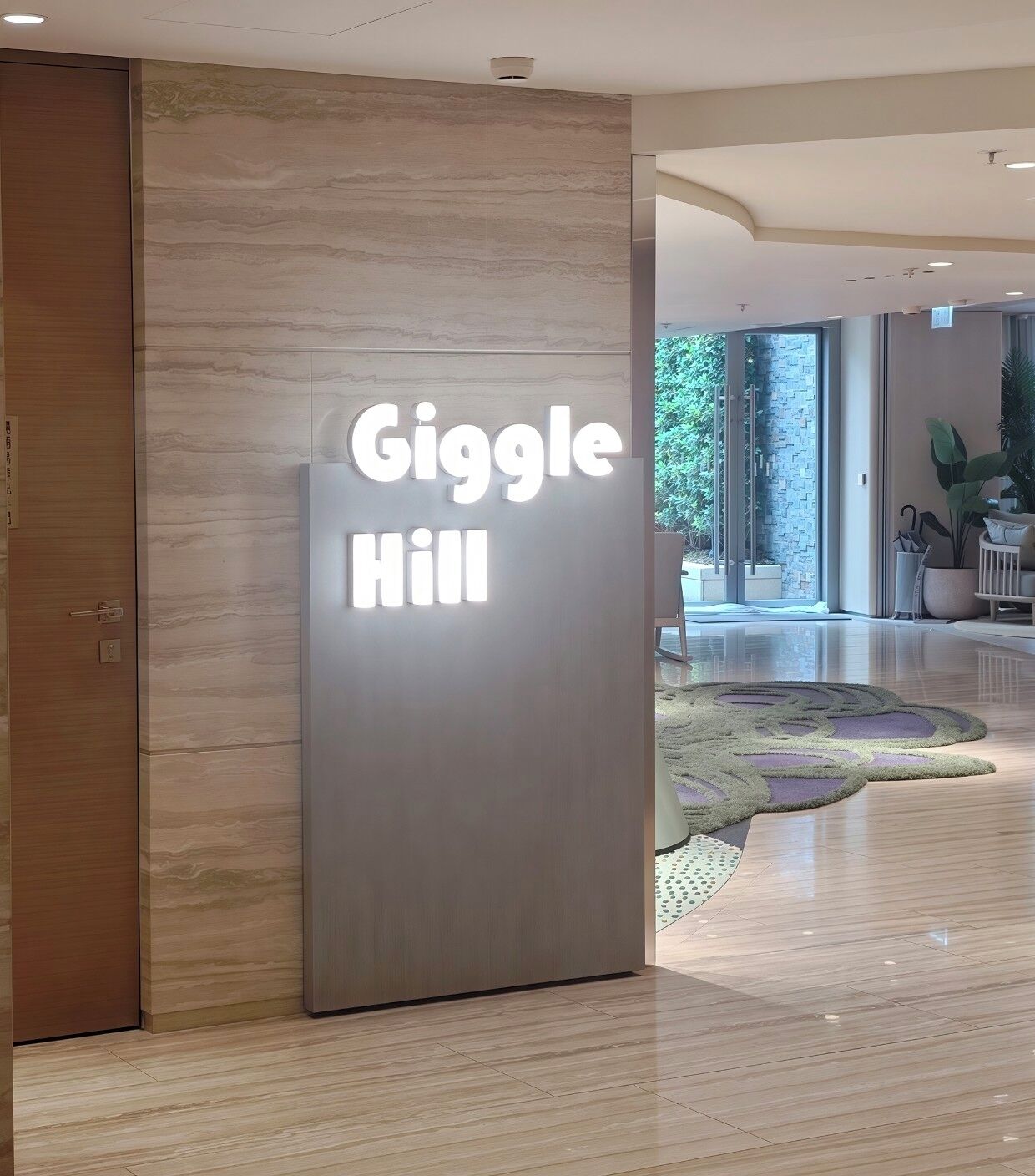 A modern hotel entrance with a 'Giggle Hill' sign, a wooden door, and a glass wall offering a view of greenery.
