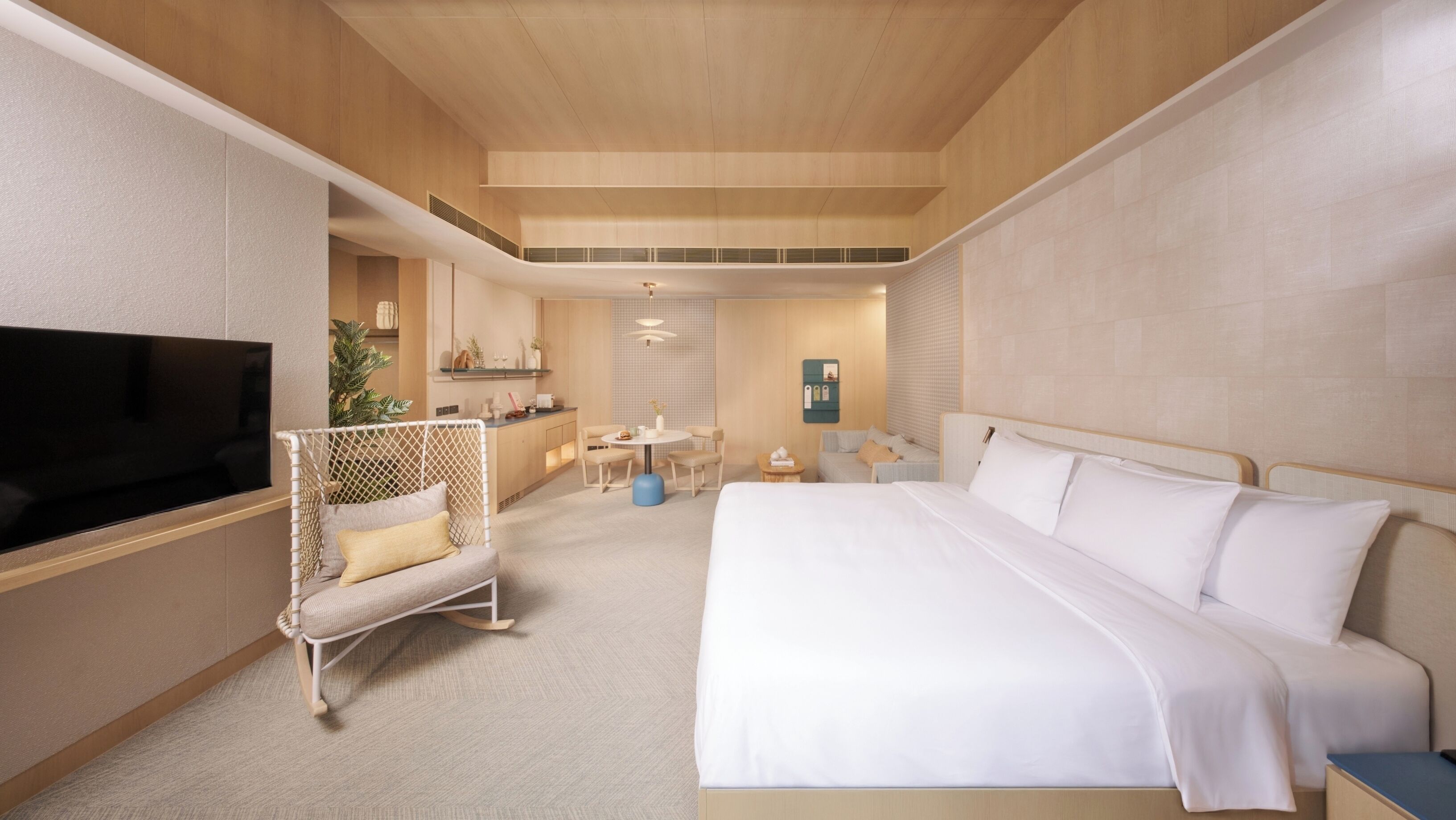 A modern hotel room with a large bed, a TV, a seating area with a plant, and a kitchenette in the background.