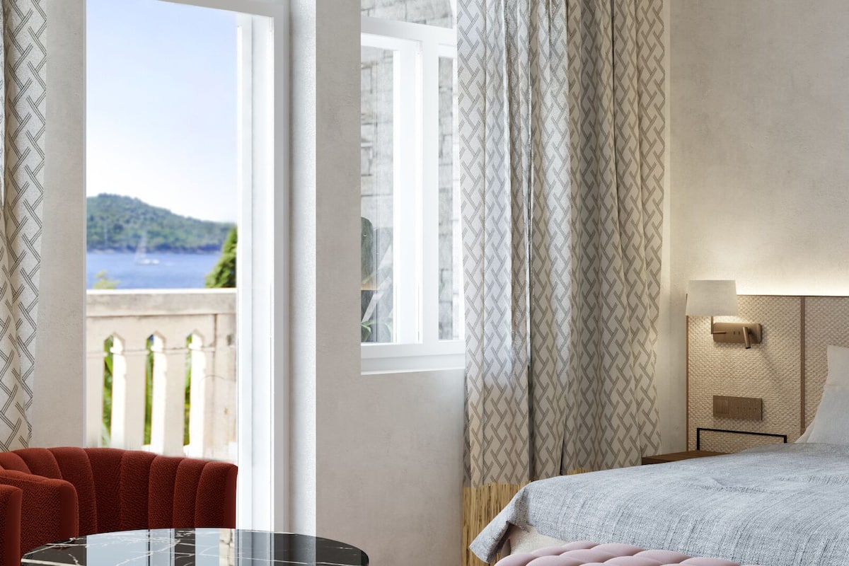 A hotel room with a bed, a small table, and a chair. There is a window with curtains and a view of a body of water and hills.