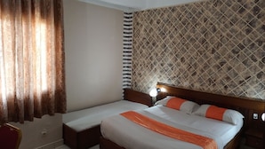 A hotel room with a bed, a bedside table, a chair, and a patterned wall. - Hotel Jouvence 2000 (Yaoundé)