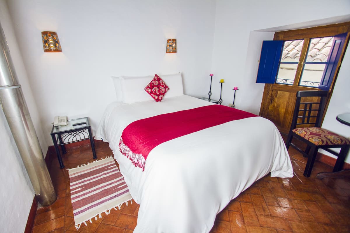 A hotel room with a bed, a nightstand, a chair, a wooden door, and a window with blue shutters.