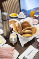 Een ontbijttafel met pannenkoeken, ham, een thermoskan en een fruitschaal.