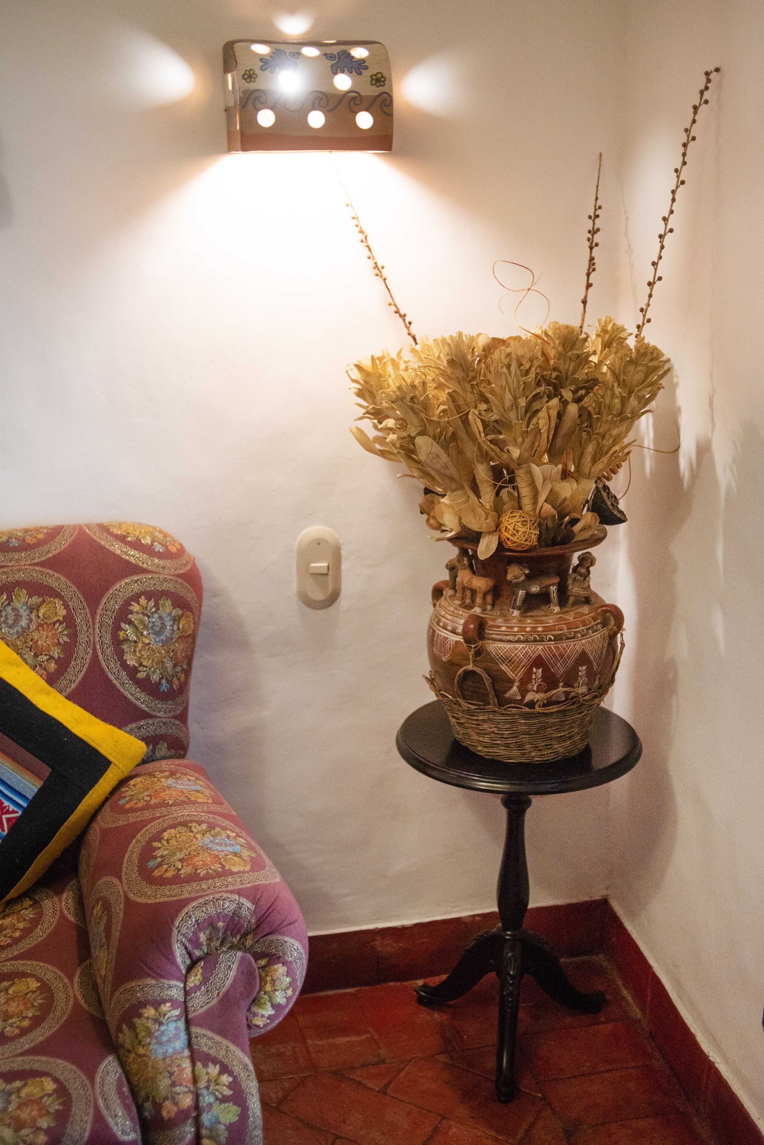 Un rincón con un sofá estampado, un jarrón decorativo con flores secas y una lámpara de pared.
