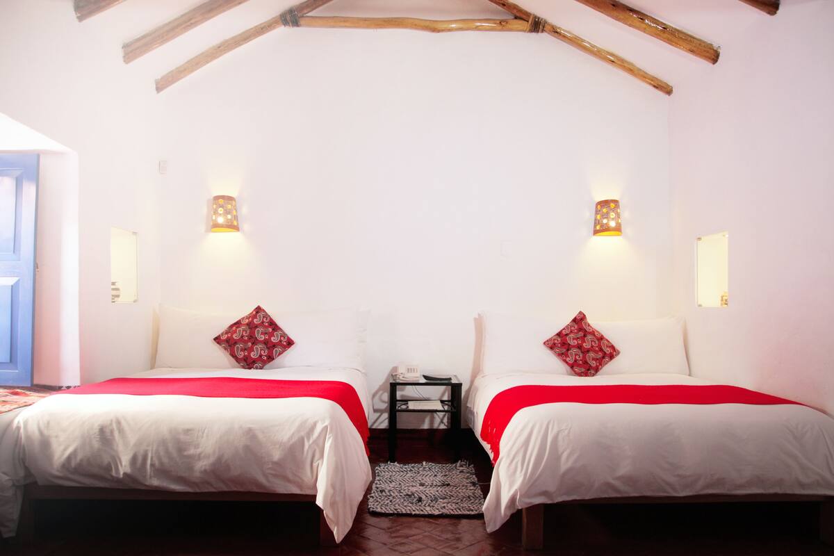 A room with two beds, each with a red and white patterned pillow, white bedding, and a small table between them.