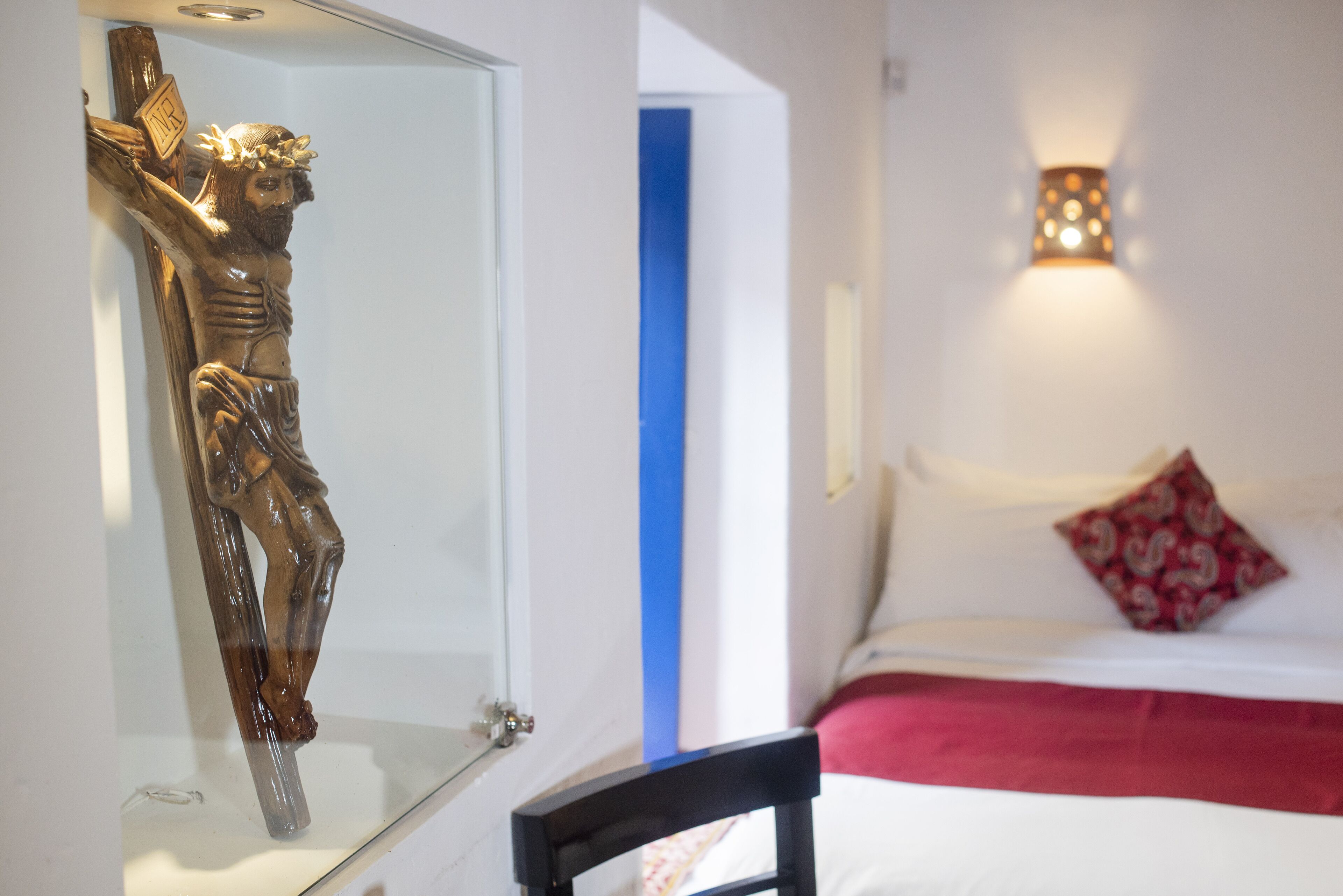 Una habitación con un crucifijo de madera sobre un expositor de vidrio, una cama con cubrecolcha rojo y blanco, y un aplique de pared.