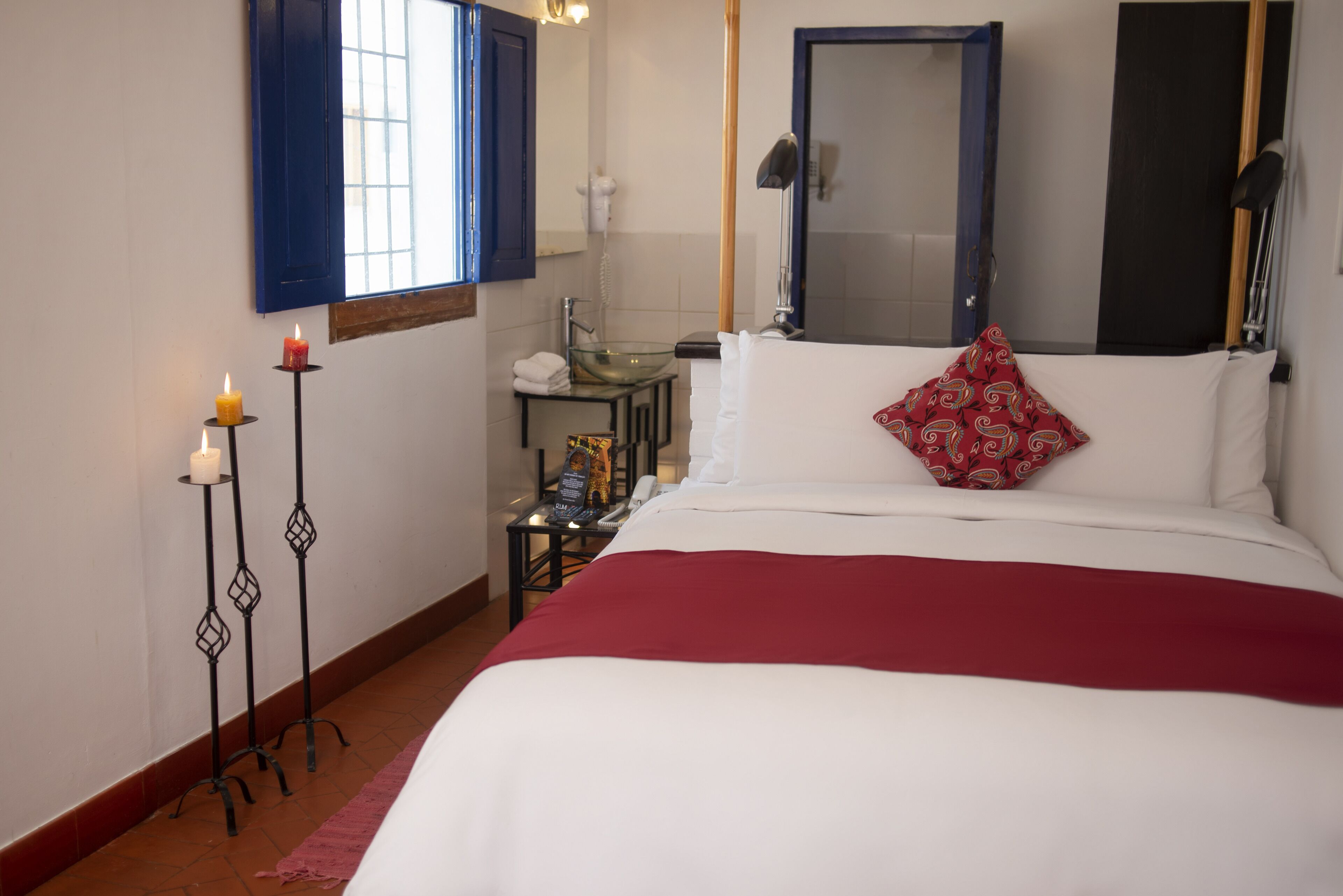 A hotel room with a bed, a nightstand, a mirror, a window with blue shutters, and a bathroom visible in the background.