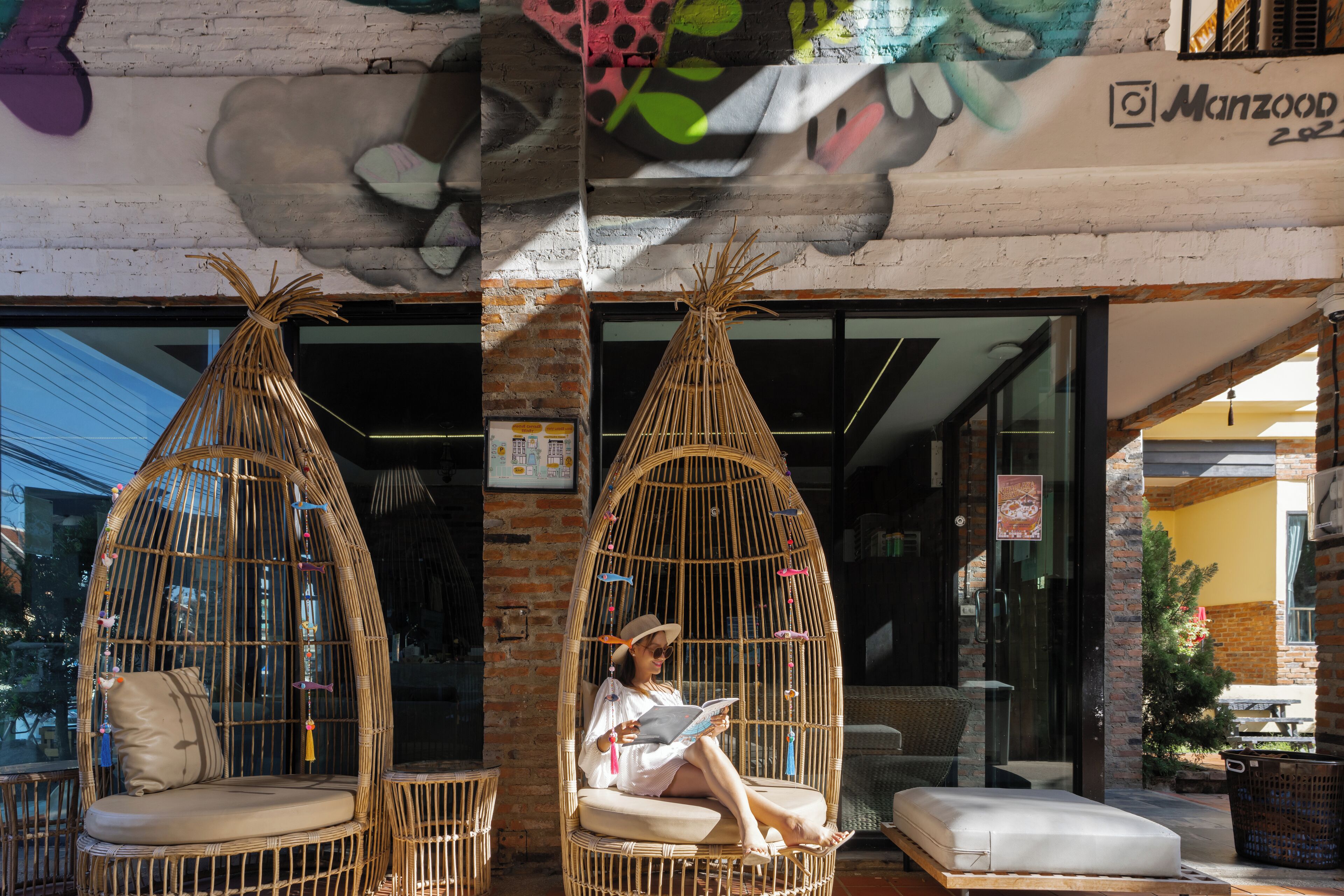 Una persona sentada en una silla de mimbre con cojín, leyendo un libro, frente a un edificio de fachada de ladrillo y entrada de vidrio.