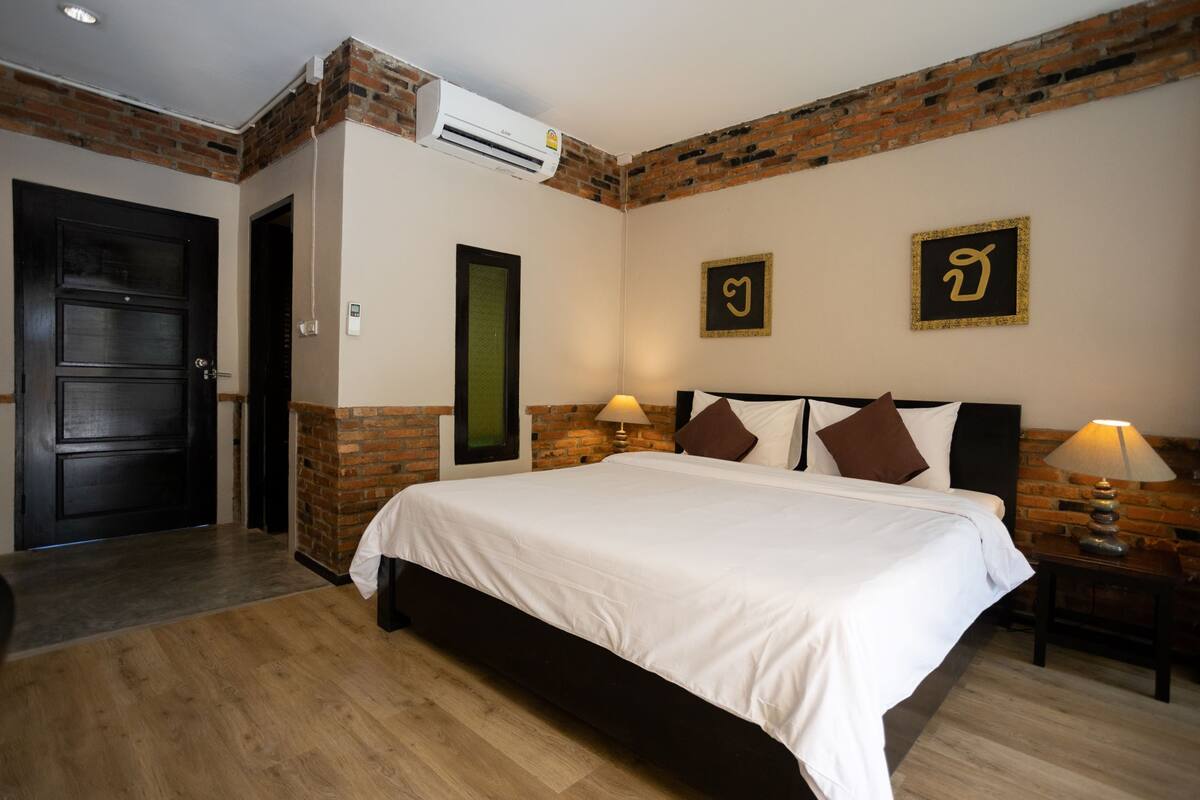 A bedroom with a brick accent wall, a large bed with white bedding, two bedside lamps, and a door with a green glass panel.