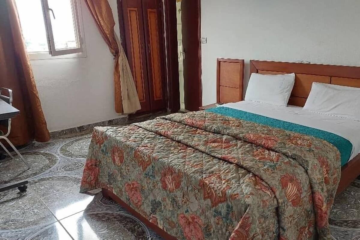 A bedroom with a bed, a wooden headboard, a patterned bedspread, and a window with curtains.