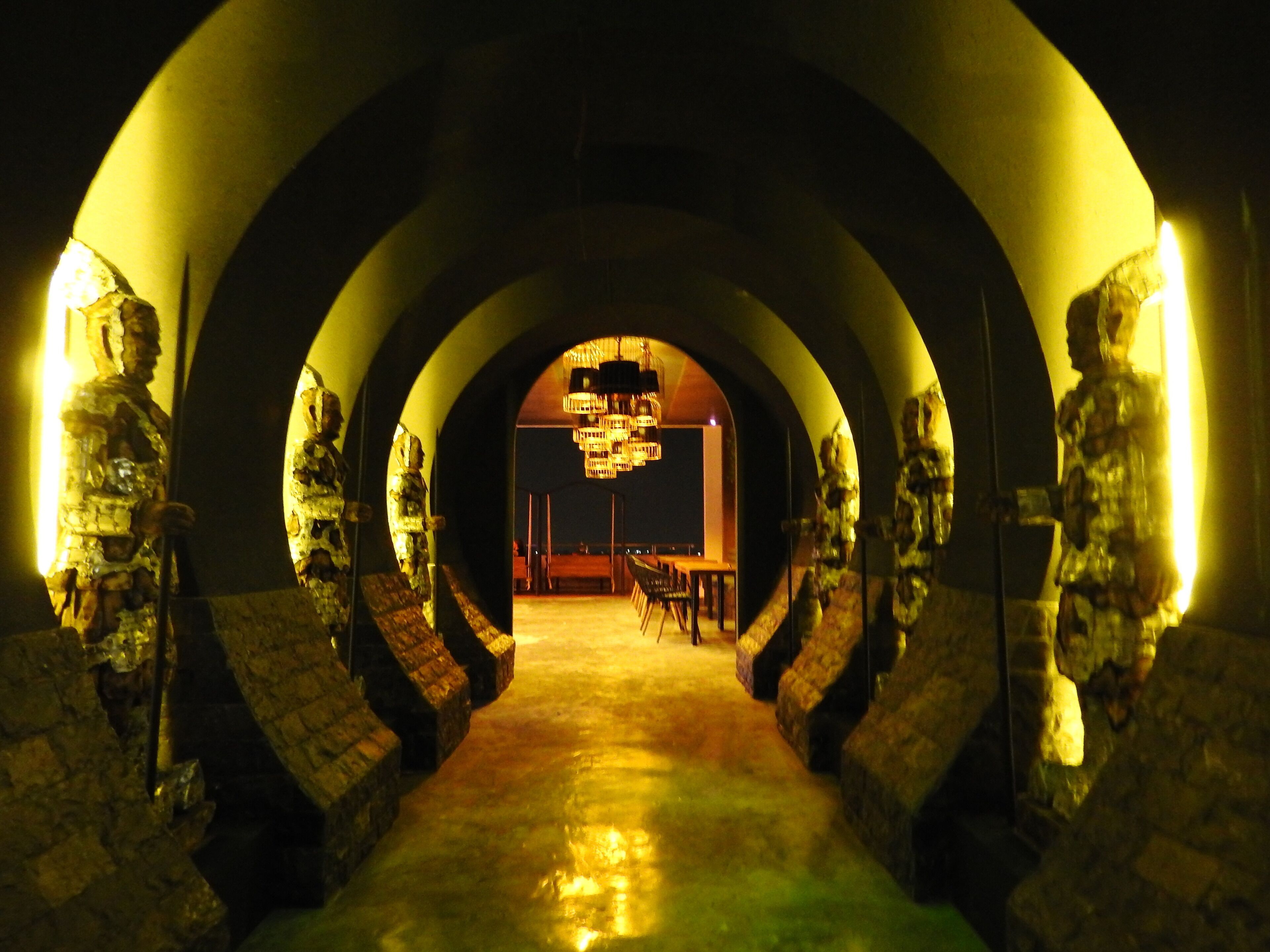 Un pasillo flanqueado por estatuas de piedra que conduce a una sala con candelabro y una mesa.