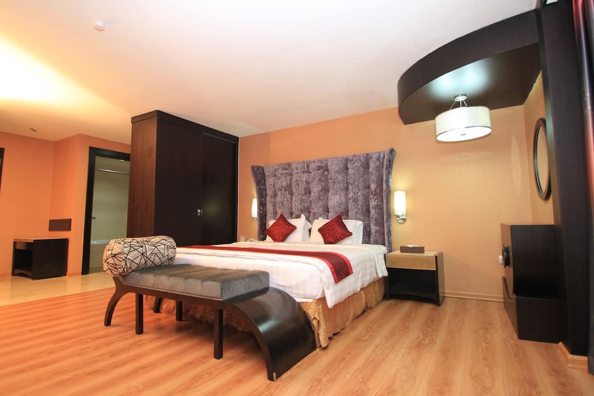 A modern hotel room with a wooden floor, a large bed with red and white bedding, a bench, a nightstand, and a wall-mounted lamp.