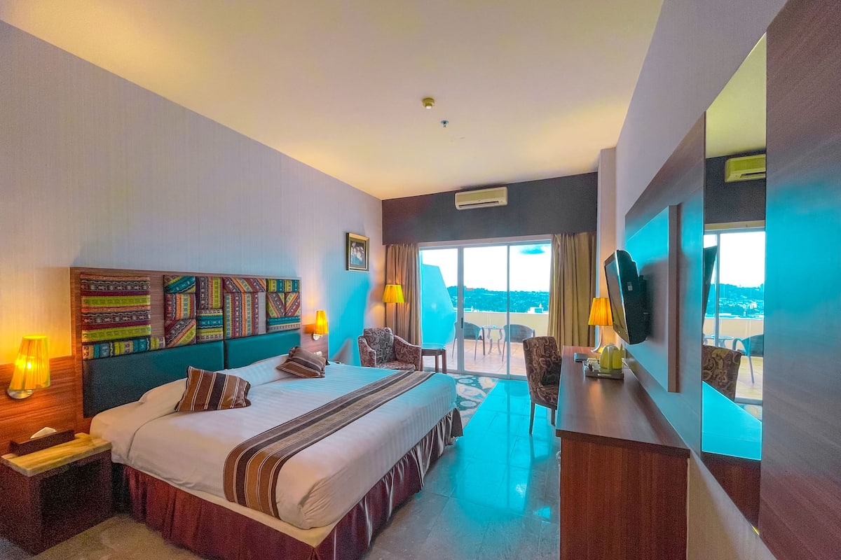 A hotel room with a large bed, a desk, a chair, a TV, and a balcony with a view.