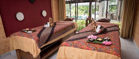 Two wooden beds with patterned bedspreads, a wooden wall decor, and a view of a garden through large windows.