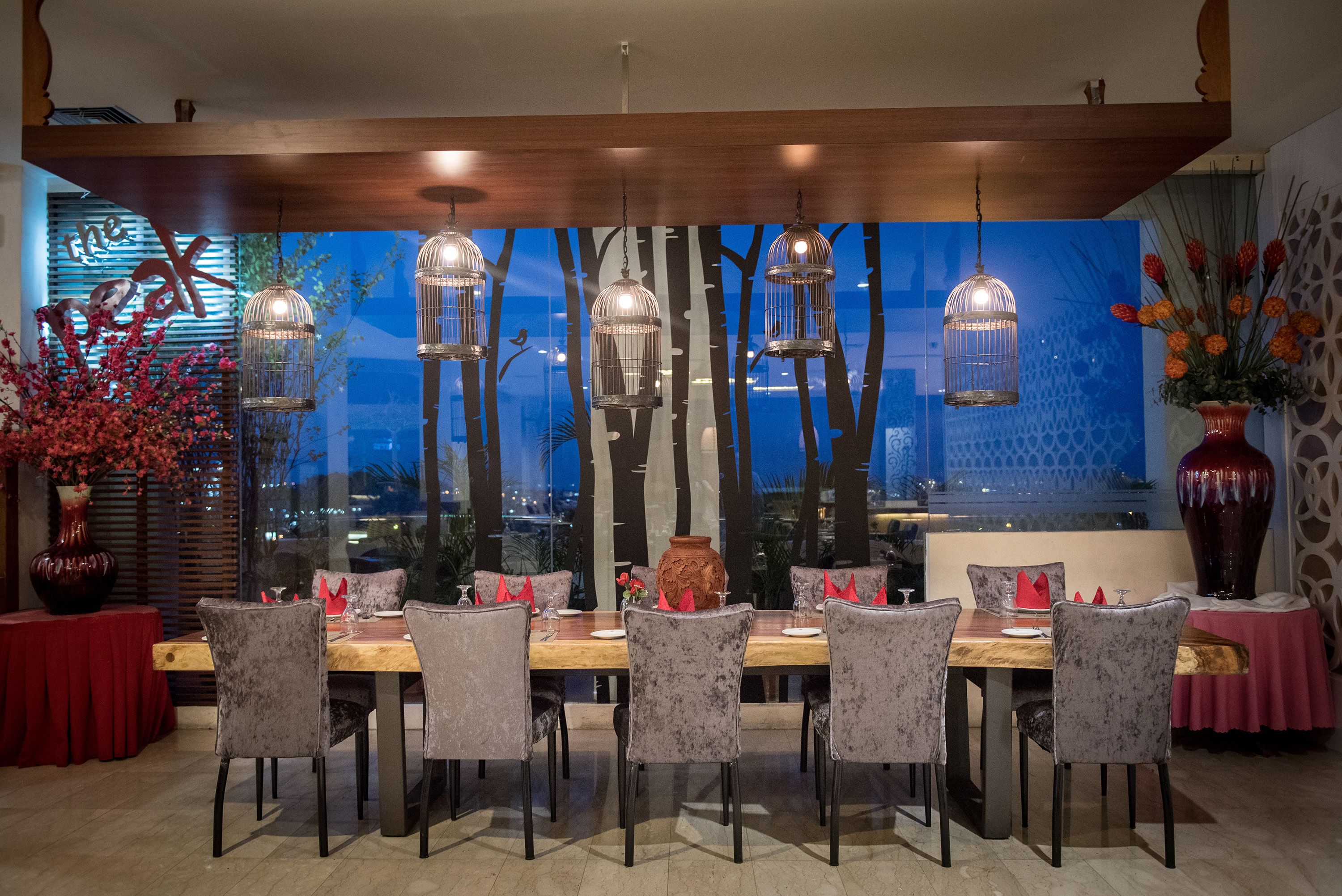 Un comedor con mesa de madera, sillas grises tapizadas, lámparas colgantes en forma de jaula de pájaros y vistas a árboles a través de una ventana.