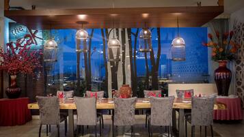 Un comedor con mesa de madera, sillas grises tapizadas, lámparas colgantes en forma de jaula de pájaros y vistas a árboles a través de una ventana.