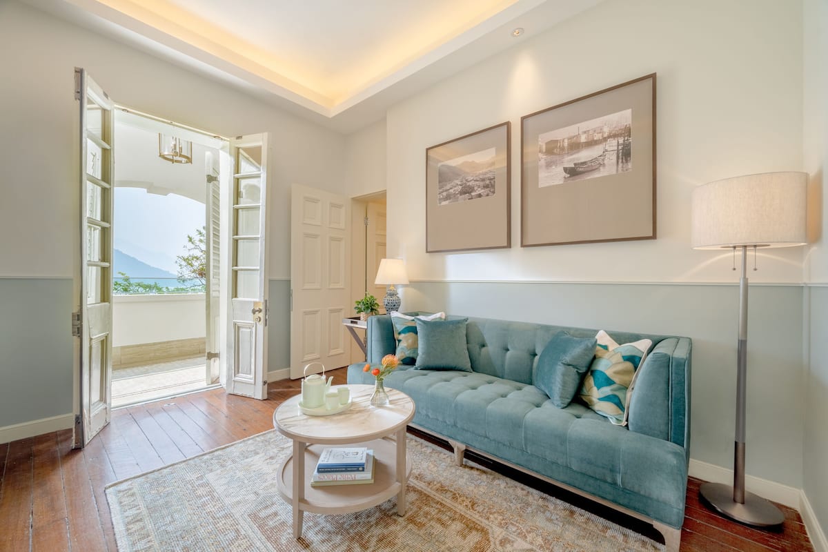 Een woonkamer met een blauwgroene bank, een ronde salontafel en een vloerlamp. Twee ingelijste foto's hangen aan de muur en er is een deur naar een balkon.