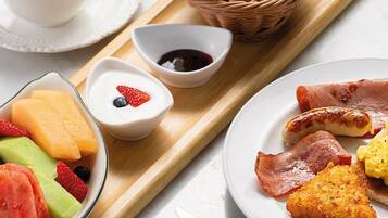 A breakfast tray with croissants, ham, sausages, fried eggs, and fruit.