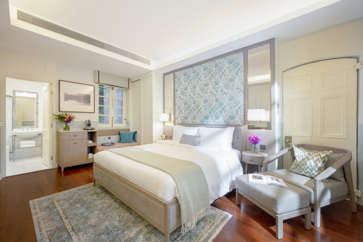 Een hotelkamer met een groot bed, een nachtkastje met een lamp, een stoel, een kleintje tafeltje met een vaas en een spiegel.