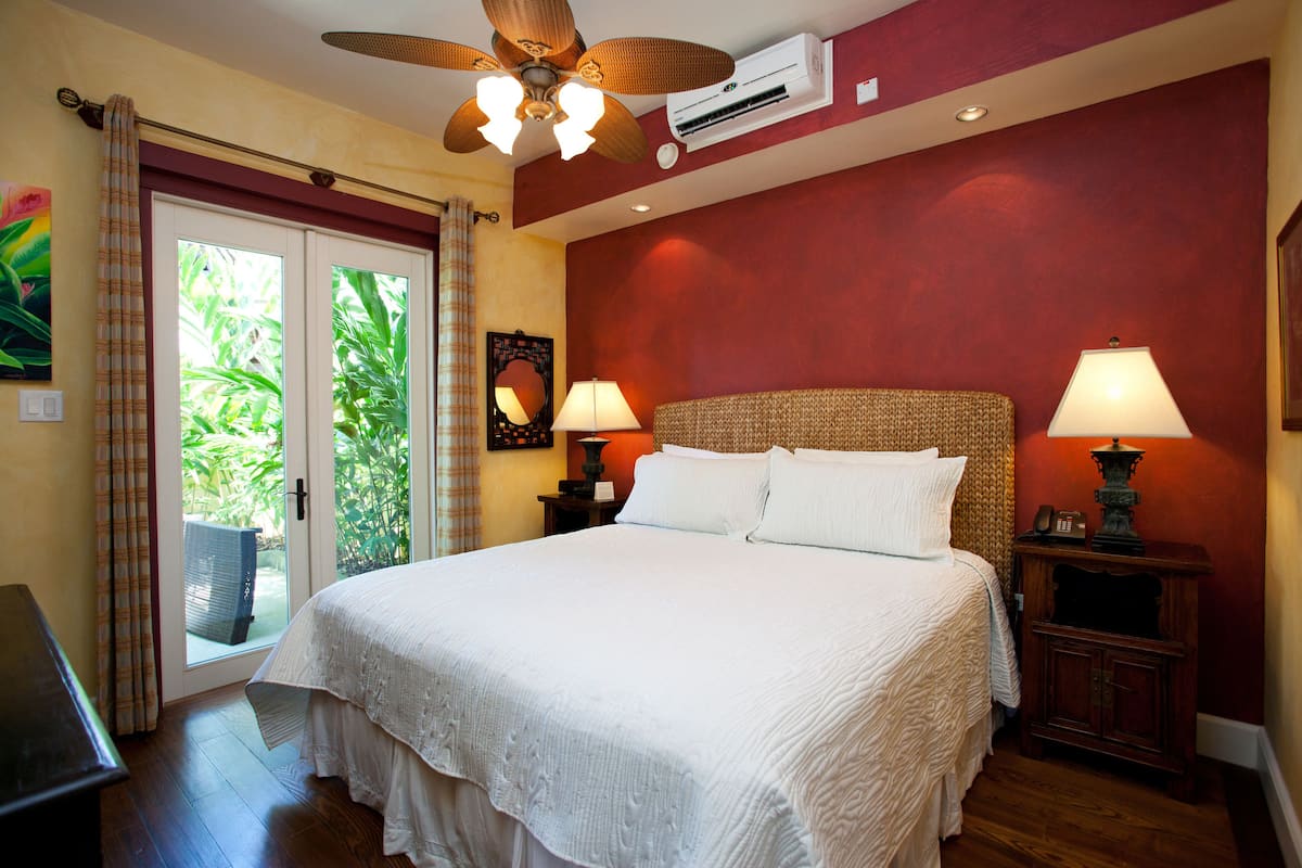 A hotel room with a large bed, two bedside lamps, a ceiling fan, and a view of greenery through the door.