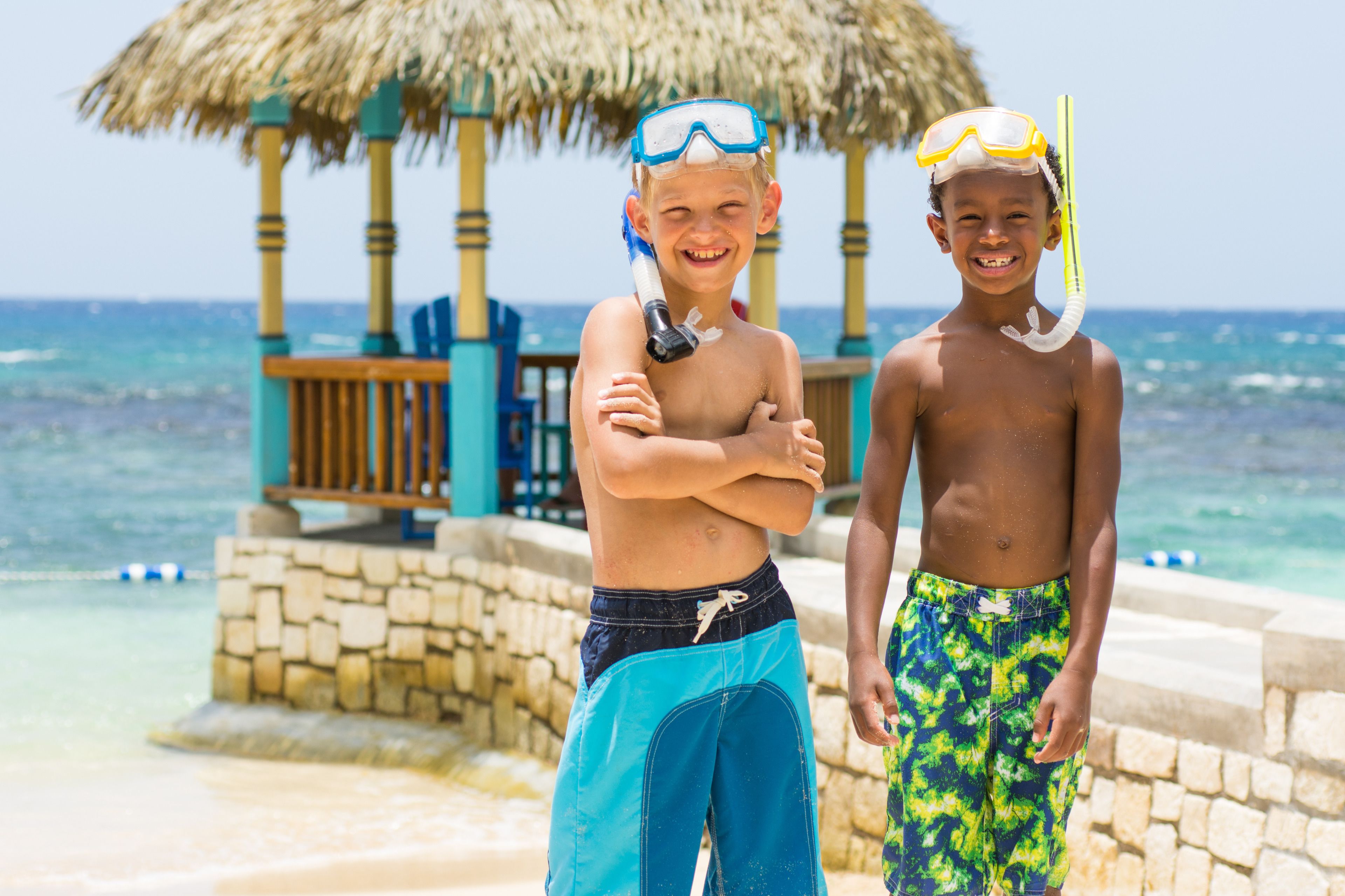 Two children snorkeling at a beach with a thatched-roof structure in the background.