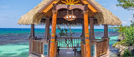 A wooden gazebo with a thatched roof overlooking a turquoise sea.