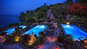 Ein Resort mit zwei Pools, umgeben von tropischer Vegetation und einem Gebäude mit Reetdach.